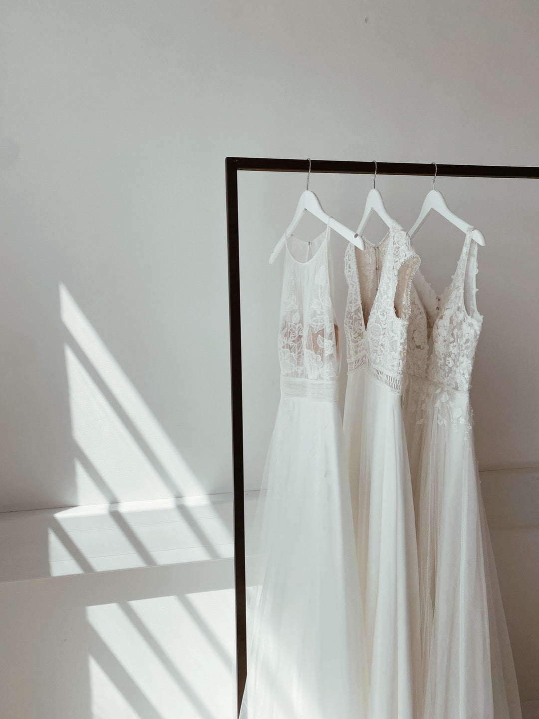 A rack of three sleeveless white Kelsey Rose wedding dresses in a room with sunshine bouncing off the white wall