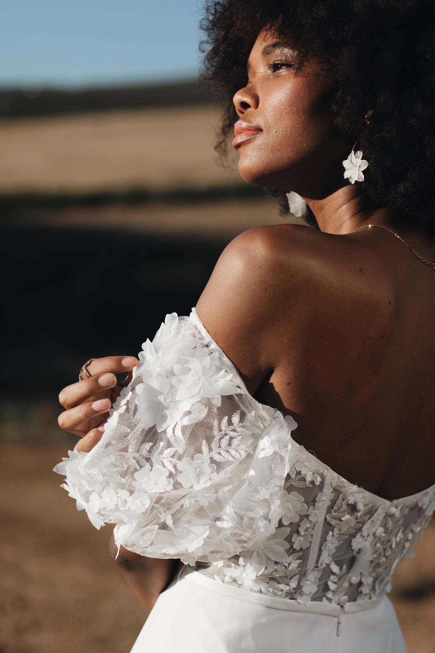 A woman with black curly hair is looking in the distance. The woman is wearing a drop shoulder floral bridal top and a white skirt, both designed by Kelsey Rose Bridal