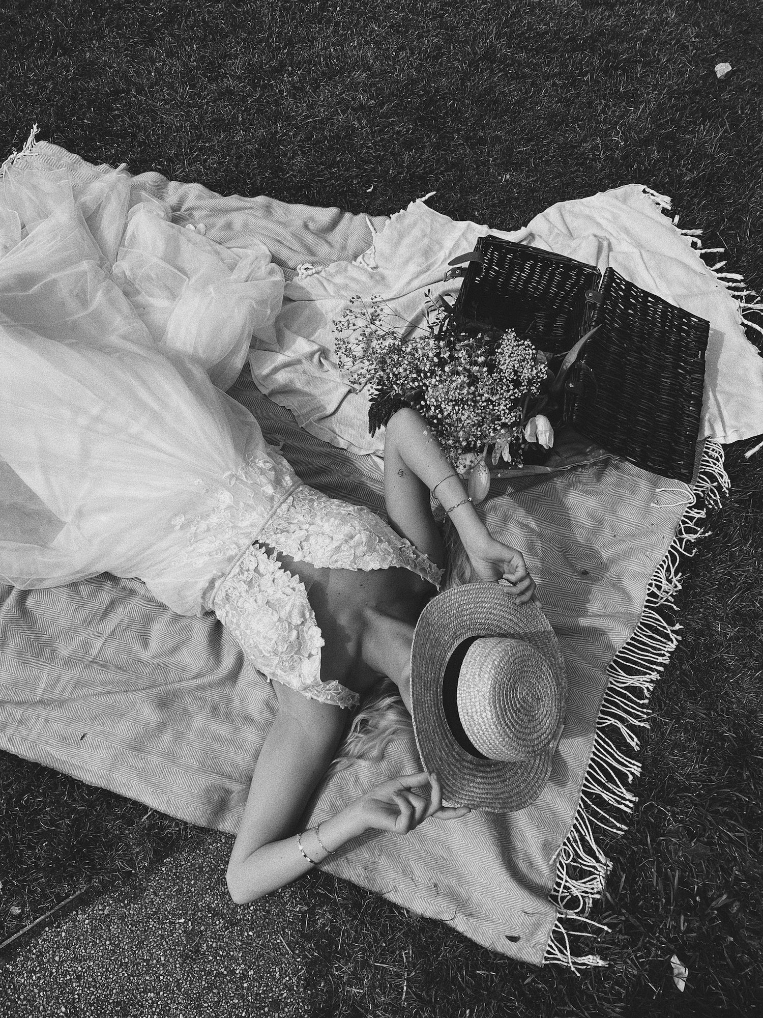 A black and white image of a woman laying on a blanket on the grass and to the left of her there is a picnic basket. The woman is wearing a straw hat and a wedding dress by Kelsey Rose Bridal