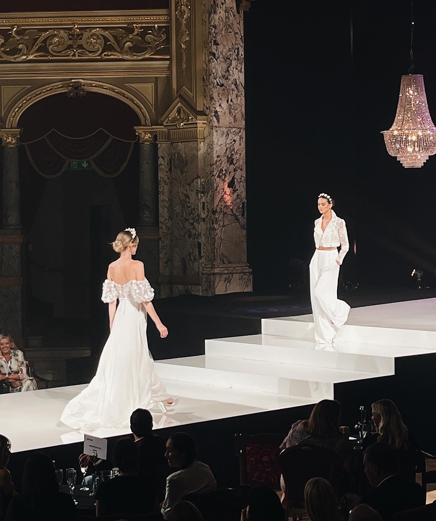 Two women are walking a white catwalk surrounded by people and photographers. One woman is wearing a white bridal jacket and long white trousers from the Kelsey Rose Bridal range. The other woman is wearing a white off the shoulder long wedding dress also from the Kelsey Rose Bridal collection
