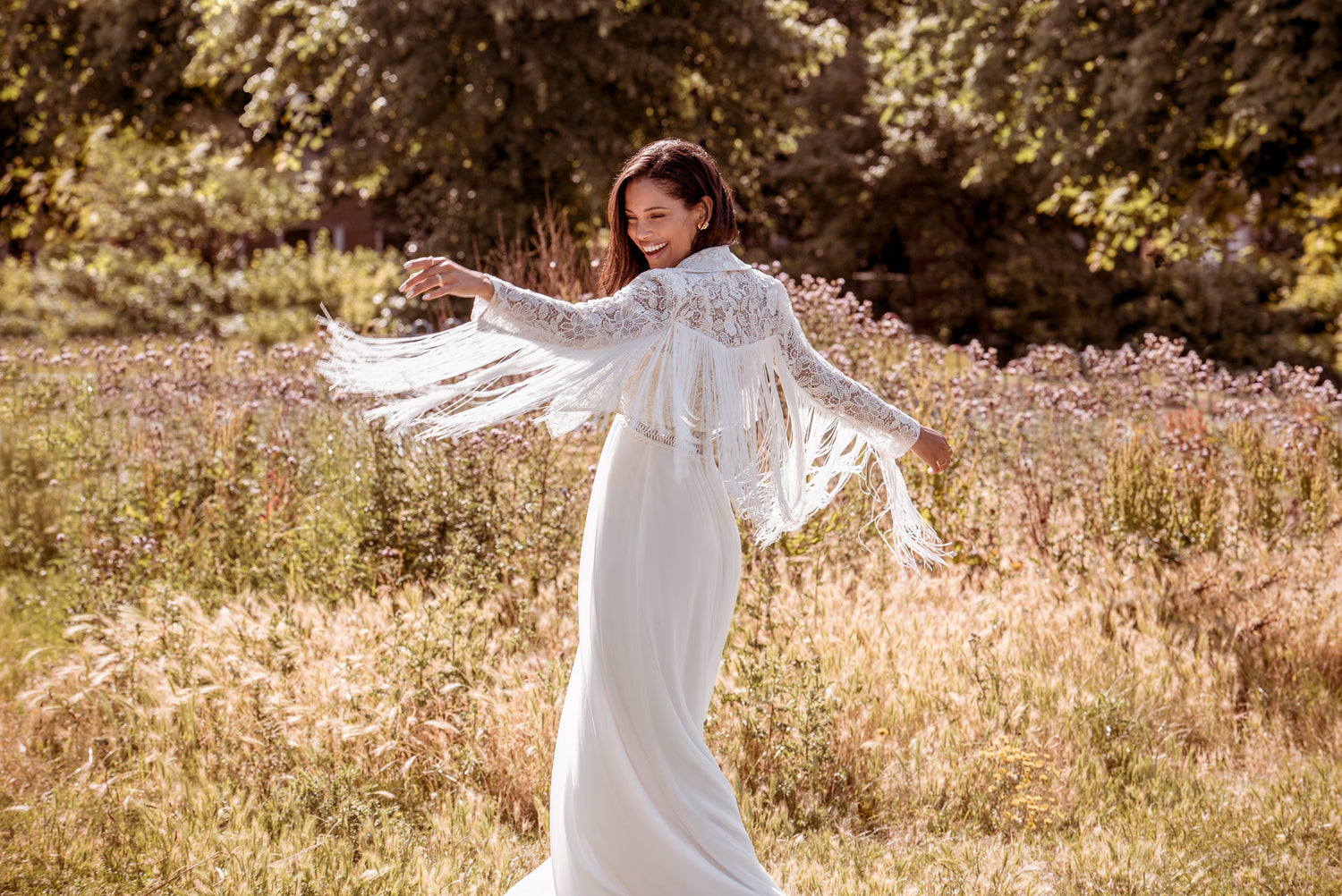A woman with dark brown hair is in the middle of a meadow. The woman is wearing the white Raine fringe jacket and a long bridal skirt. Her arms are in the air and the fringe of the jacket is flowing underneath her arms