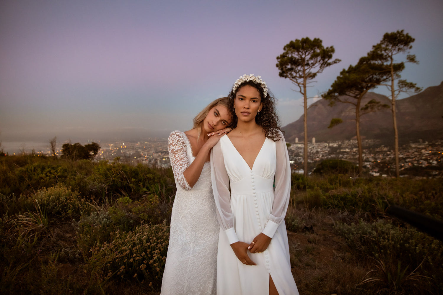 Two women surrounded by greenery and hills above the city as the sun goes down. One woman has blonde hair and is wearing a modern, long floral, three quarter sleeve dress she is resting her head and hands on the left shoulder of the other woman. The other woman has brown curly hair and is wearing a floral head piece. Her dress is long, with a deep V neck, long sheer see though sleeves and buttoned at the front..